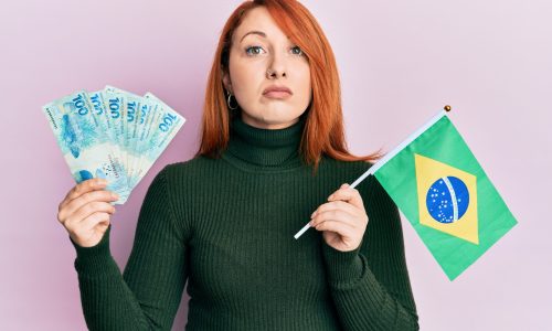 Beautiful redhead woman holding 100 brazilian real banknotes and brazil flag relaxed with serious expression on face. simple and natural looking at the camera.